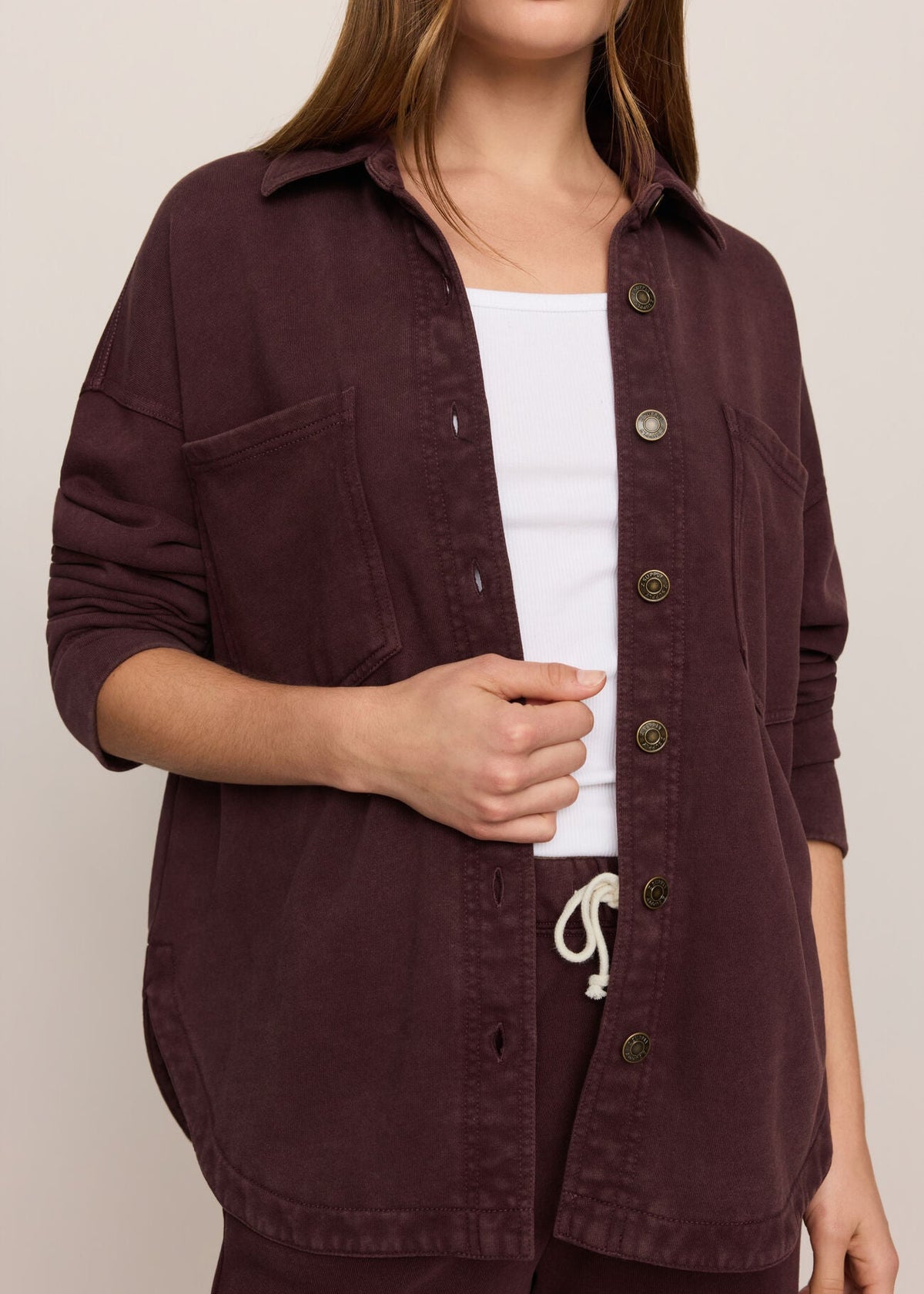A woman wearing a dark plum button-up overshirt over a white tank top, shown from the nose down against a plain light beige studio background.