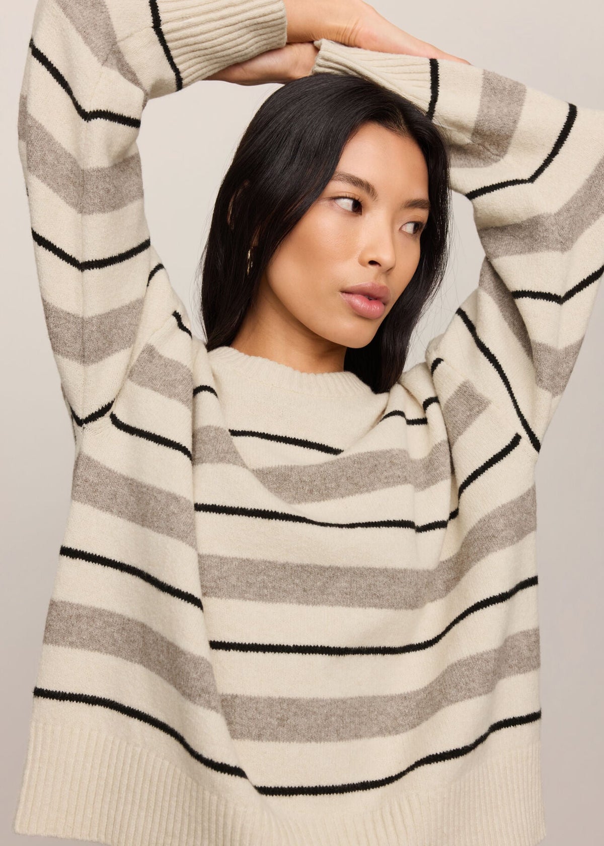 A woman wearing a cream and gray striped knit sweater with black horizontal stripes, posing with her arms raised against a plain light gray background.