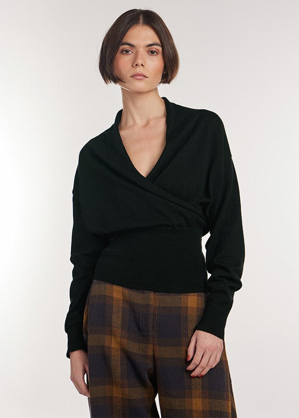 A model wearing a black long-sleeve wrap-style top and brown and black plaid trousers, standing against a plain light gray studio background.