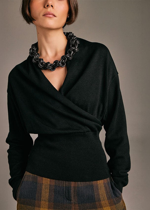 A model wearing a black knit wrap-style sweater with a chunky black chain necklace, paired with a brown and navy plaid skirt, against a neutral gray studio background.