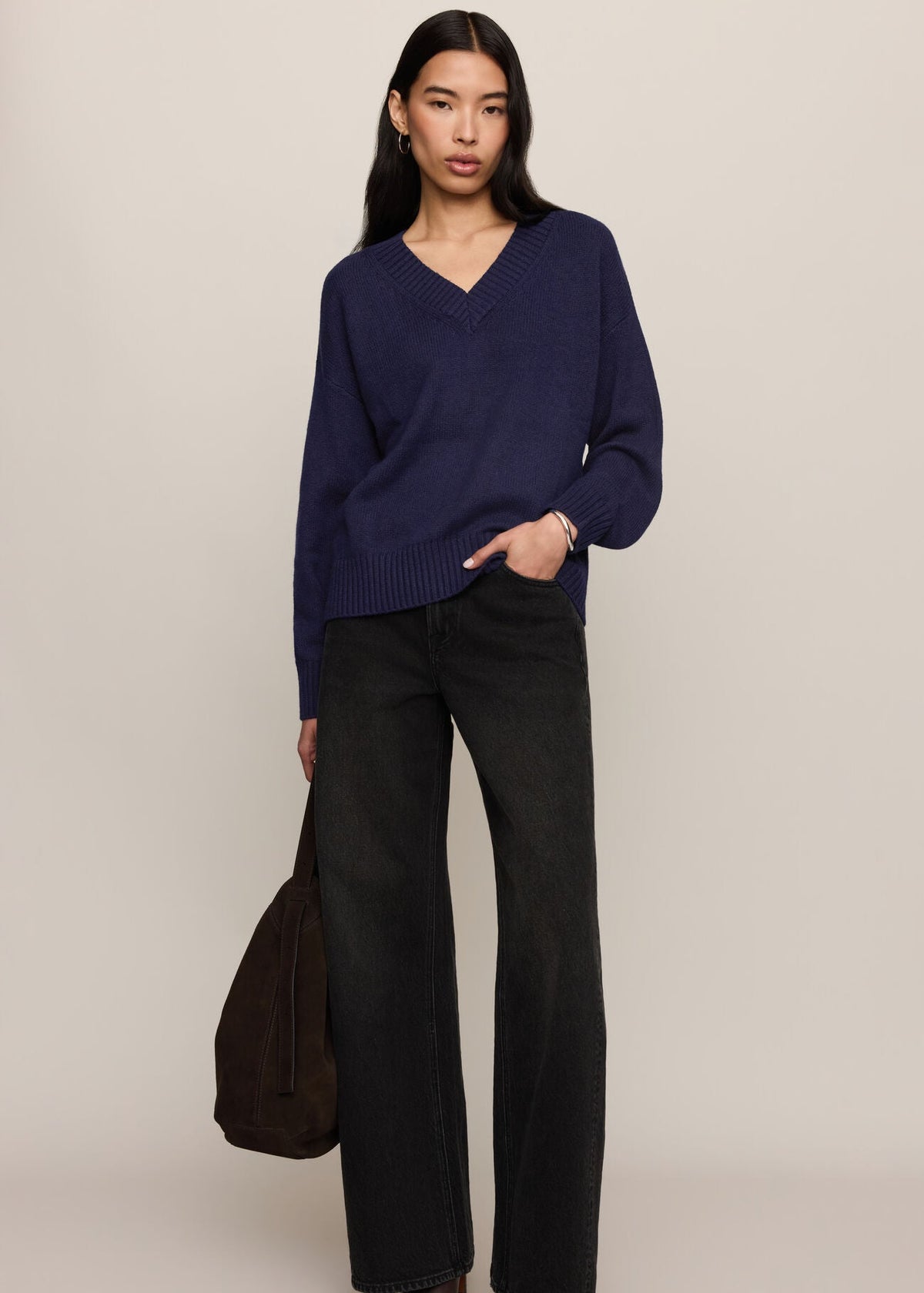 A woman stands against a plain light beige studio background wearing a navy blue V-neck knit sweater, dark wide-leg jeans, and dark pointed shoes, while holding a dark brown tote bag.