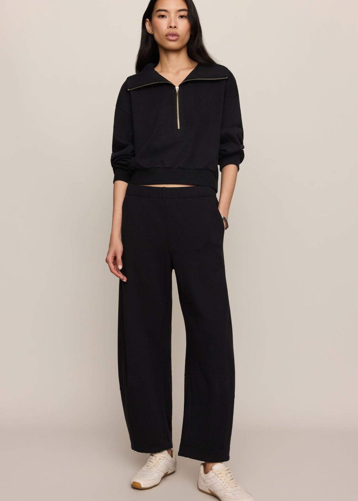 A model stands against a light beige studio background wearing a black cropped zip-up sweatshirt and matching wide-leg pants, paired with white sneakers.
