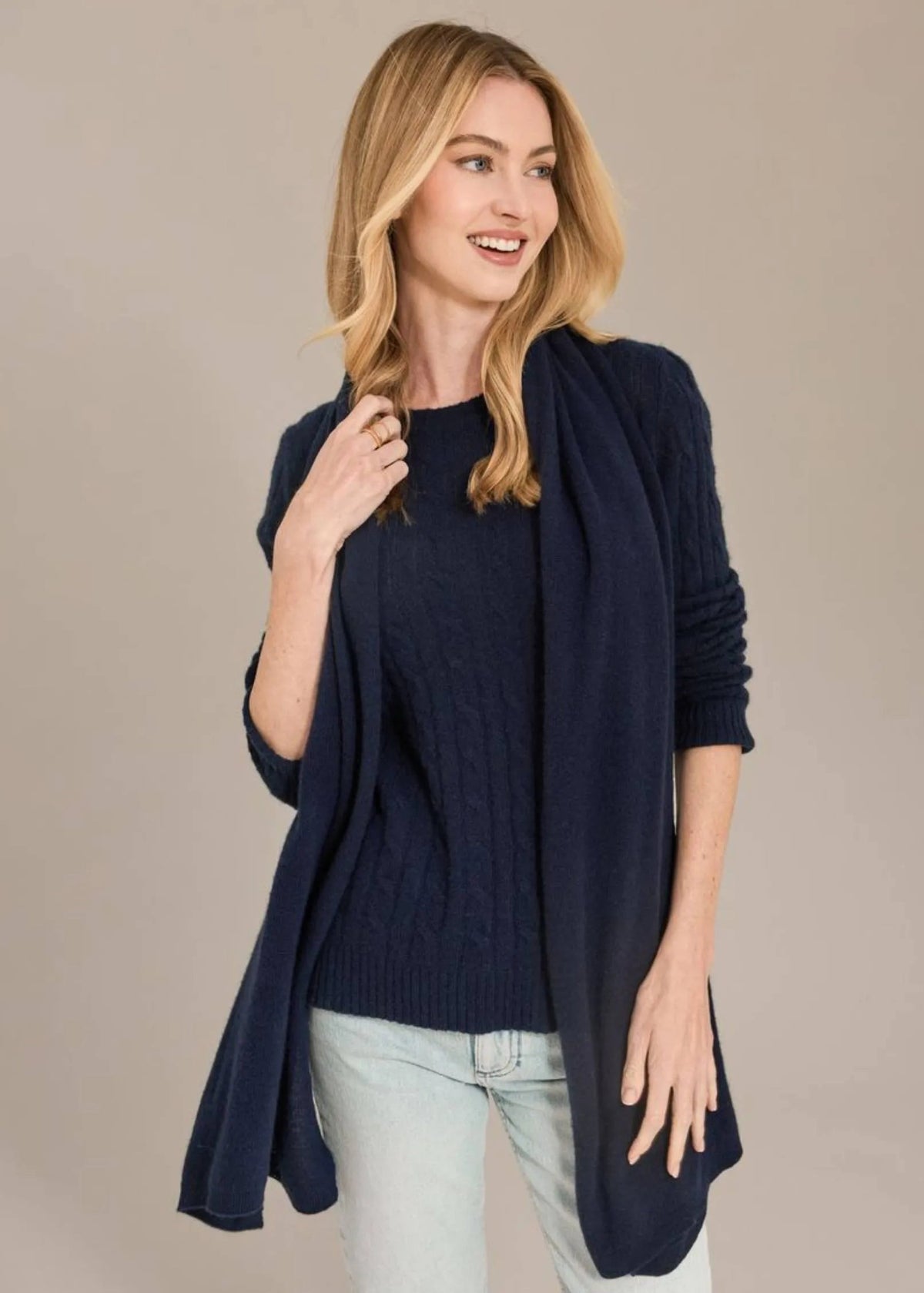 A woman modeling a navy blue cable-knit sweater layered under a matching open-front cardigan, paired with light-wash jeans, against a solid beige studio background.