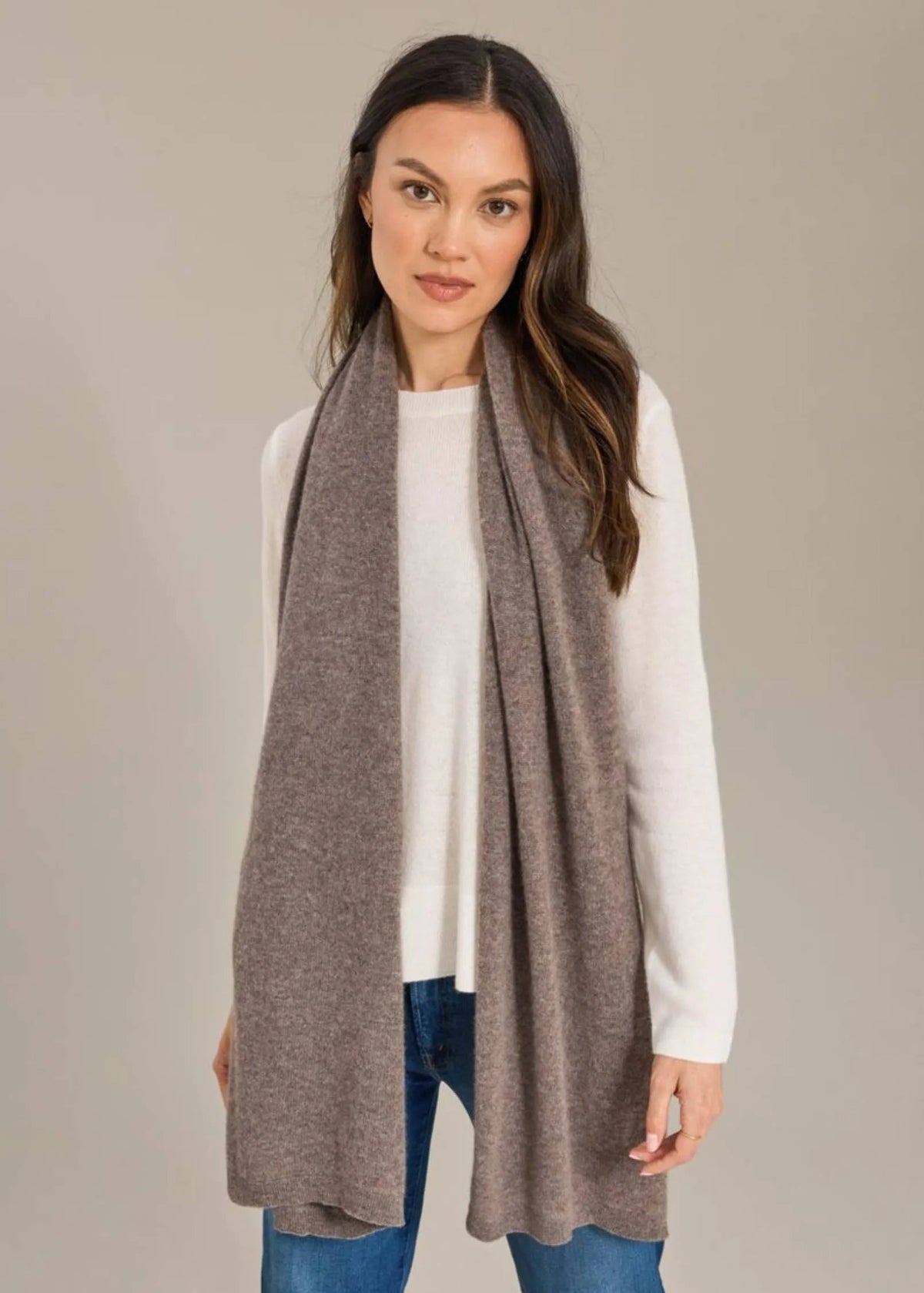 A woman modeling a long, lightweight taupe-gray knit scarf draped over a white long-sleeve top and blue jeans against a solid light gray studio background.