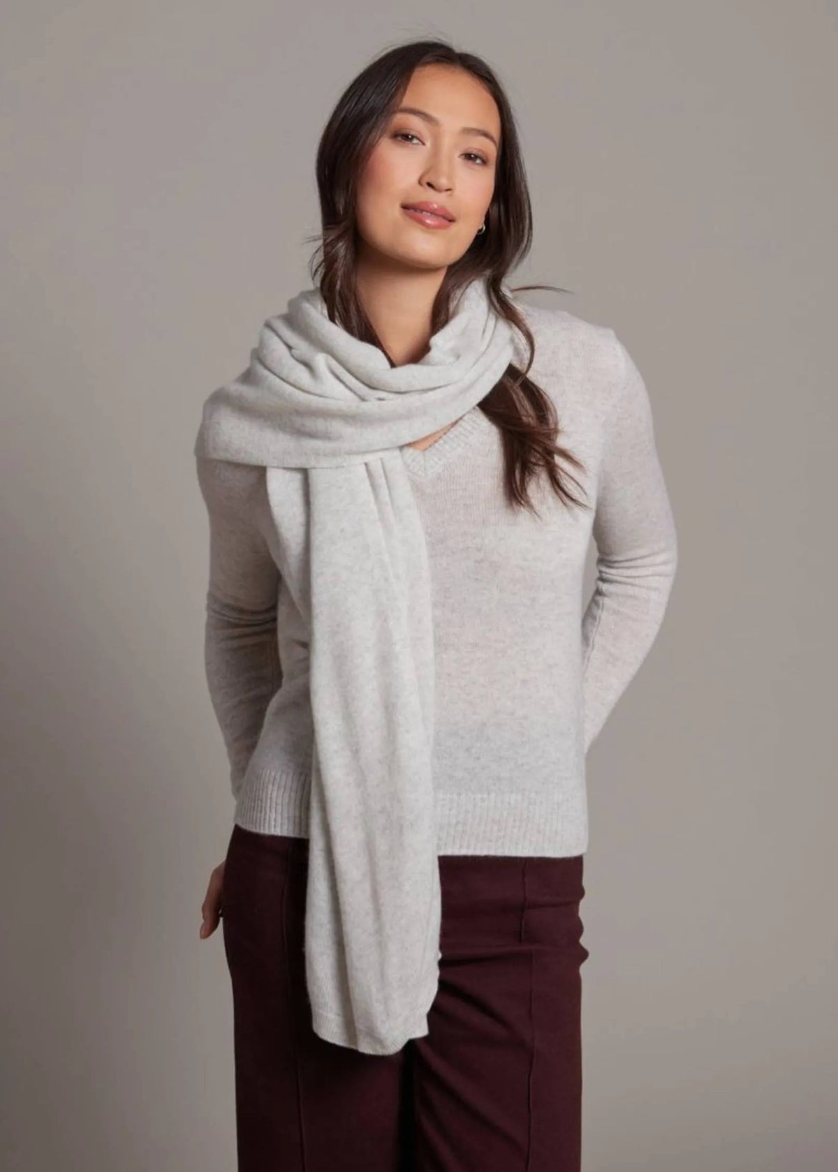 A woman modeling a light gray knit sweater and a matching light gray scarf against a plain gray studio background.