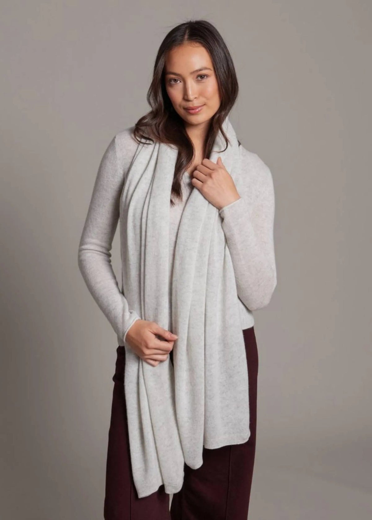 A woman modeling a light gray knit cardigan with an integrated hood and a long, draped scarf, paired with dark burgundy pants, against a solid gray studio background.