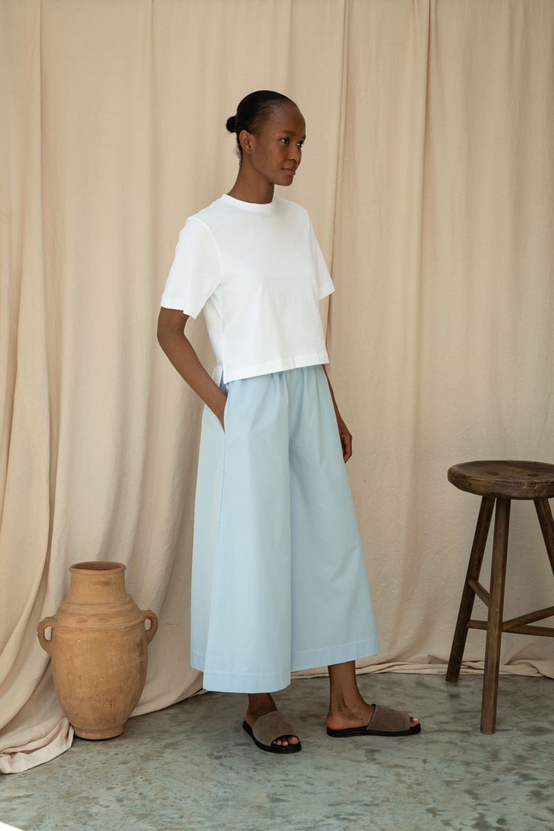 Woman wearing a white top and light blue pants standing against a beige curtain with a wooden stool and vase.