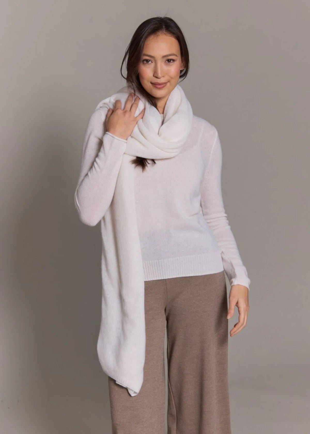 A woman modeling a light cream knit sweater and matching cream infinity scarf, paired with taupe wide-leg pants, against a neutral gray studio background.
