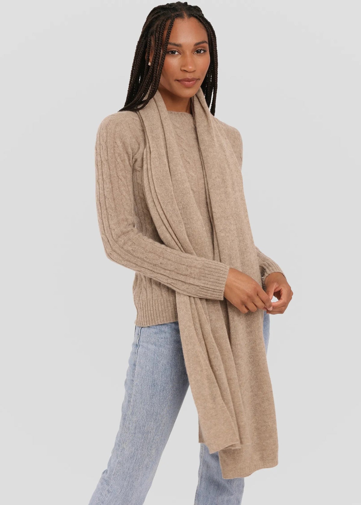 A woman modeling a long, light beige knit wrap scarf draped over a matching beige ribbed sweater, paired with light blue jeans, against a solid light gray background.