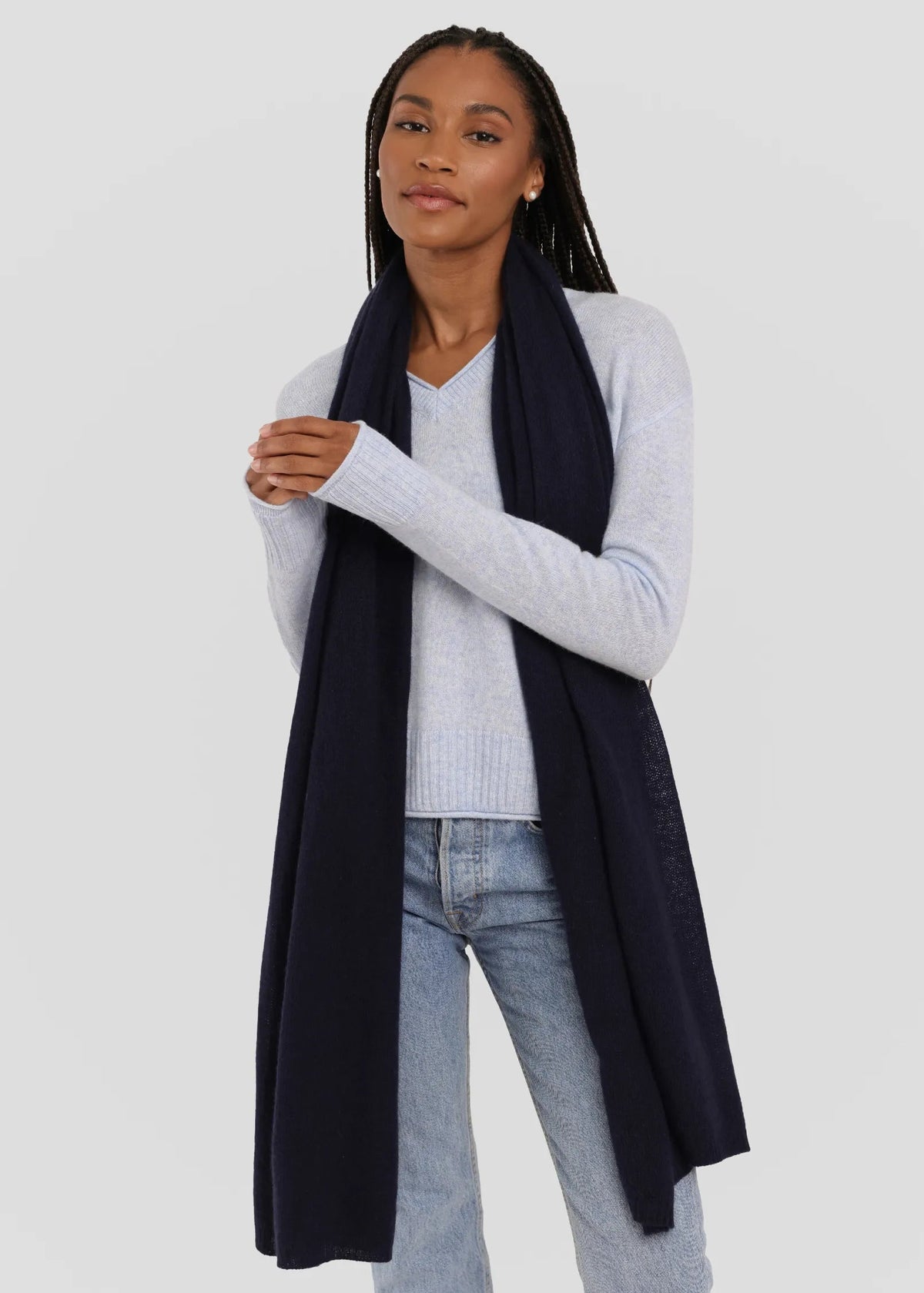 A woman modeling a long, dark navy blue knit scarf draped over a light gray sweater and light-wash jeans against a plain light gray studio background.