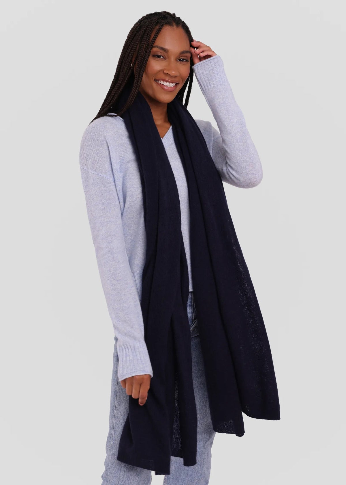 A woman modeling a long, dark navy knit scarf draped over a light gray cardigan and white top, set against a solid light gray background.