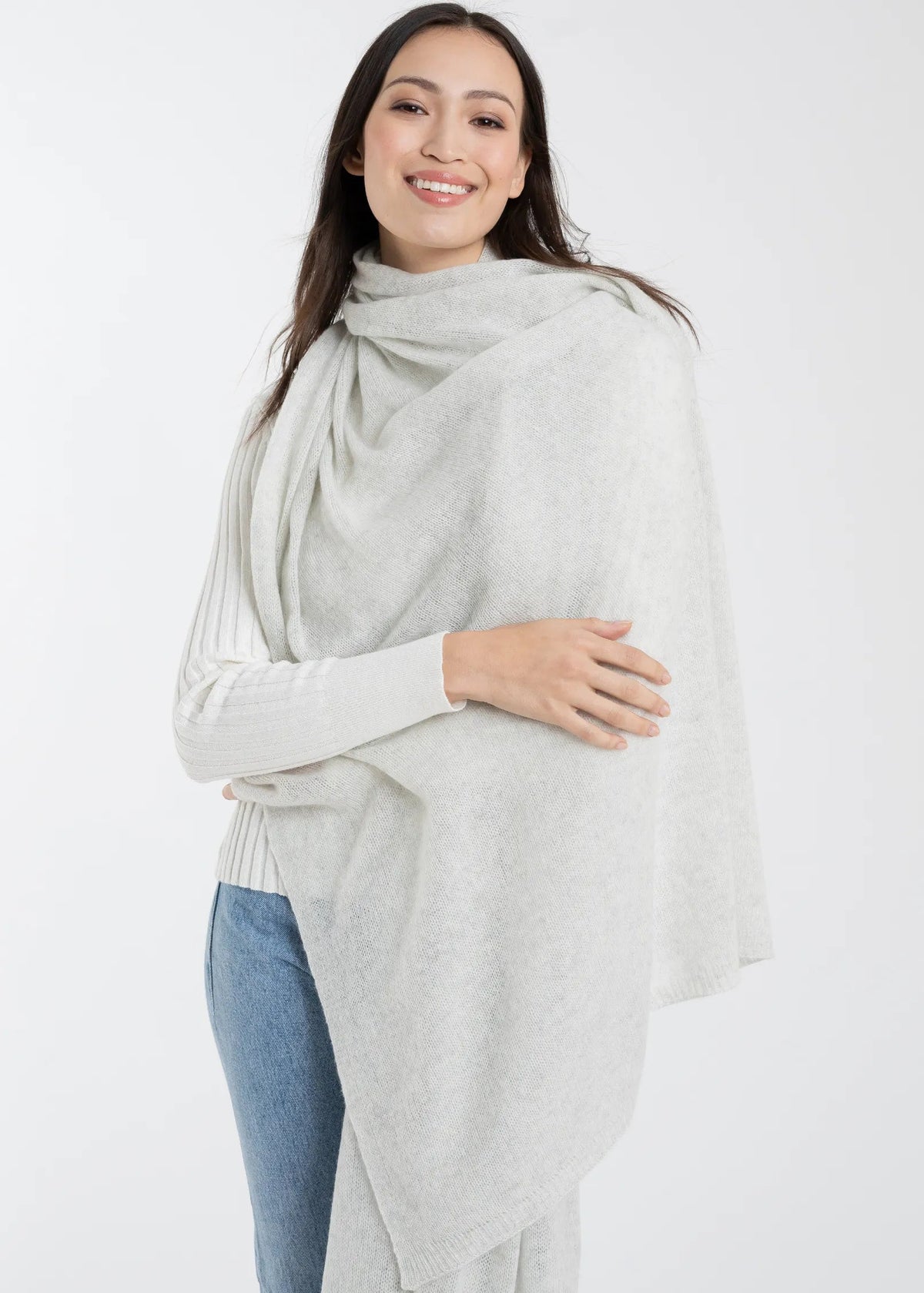 A woman modeling a light gray knit wrap or poncho draped over her shoulders, worn over a white ribbed long-sleeve top and blue jeans, against a plain white studio background.