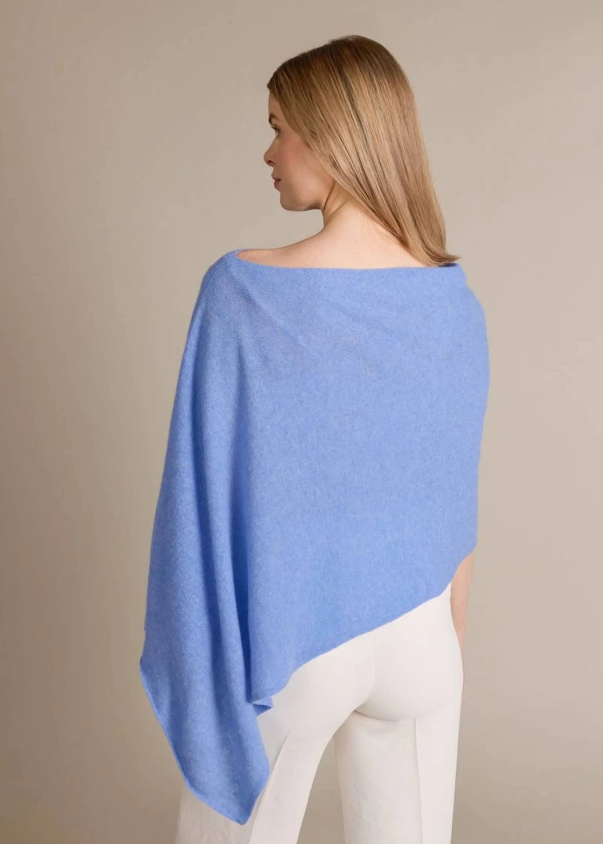 A woman shown from the back wearing a light blue draped wrap or poncho over white pants, photographed against a plain warm beige studio background.