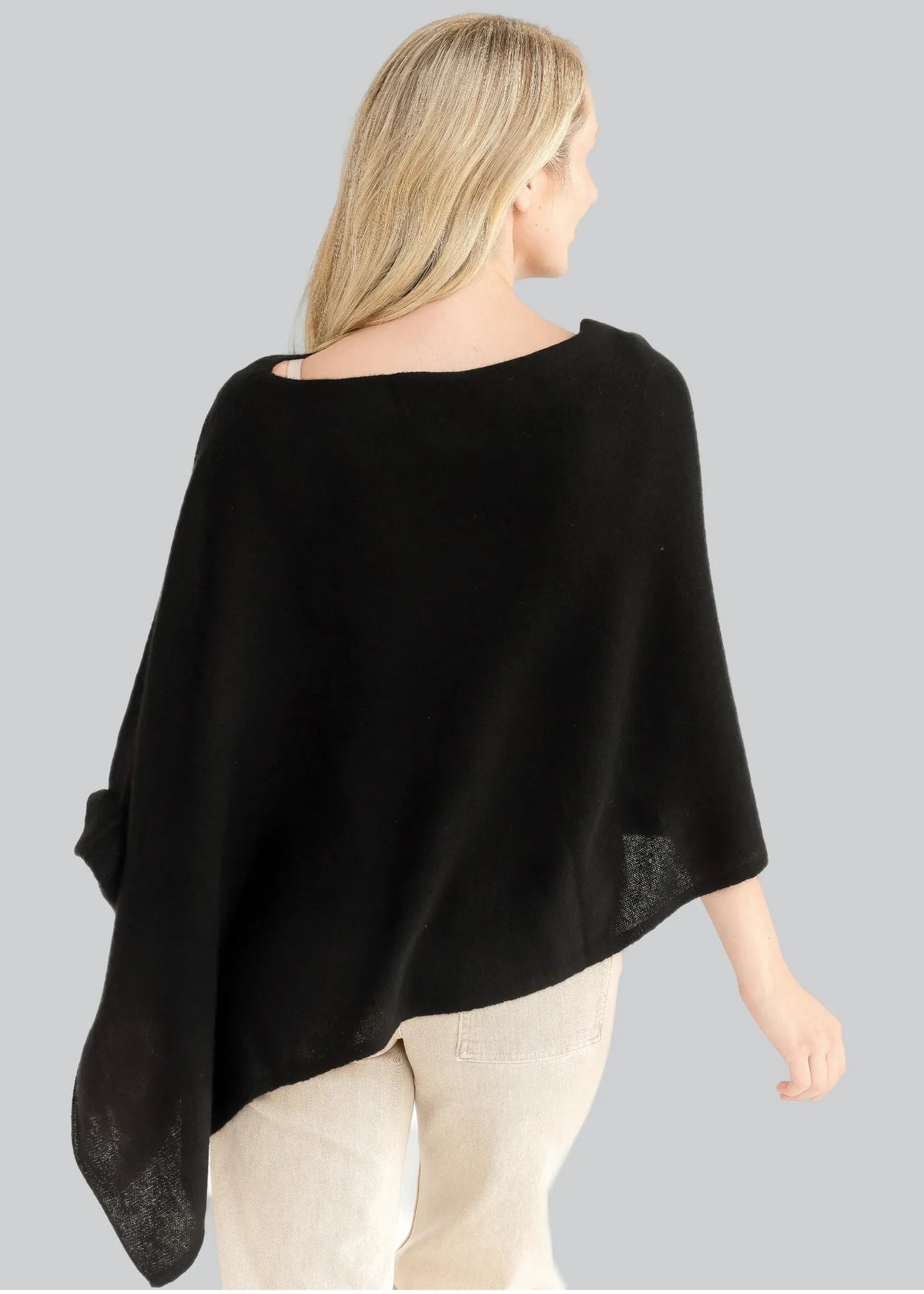 A woman shown from the back wearing a black wide-neck poncho top with textured sheer side panels, paired with light beige pants, against a solid gray background.