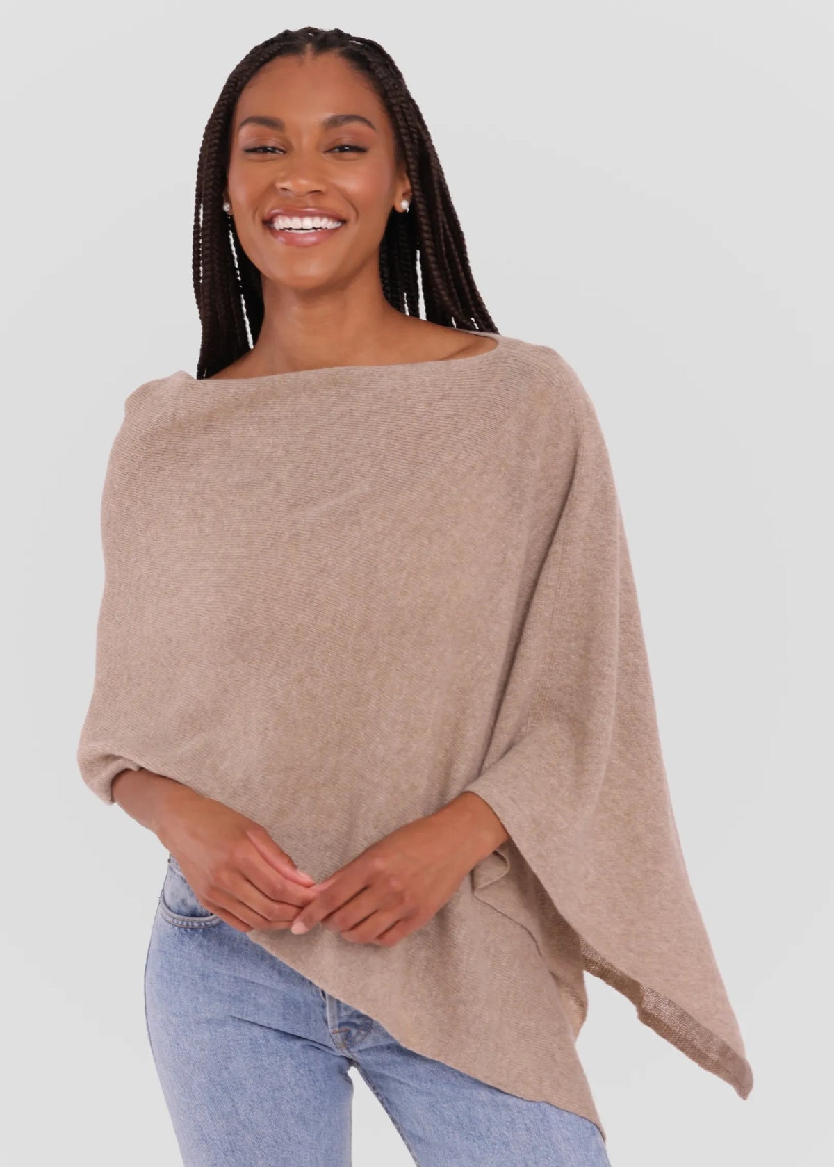 A woman wearing a light taupe knit poncho sweater and light blue jeans, standing against a solid light gray background.