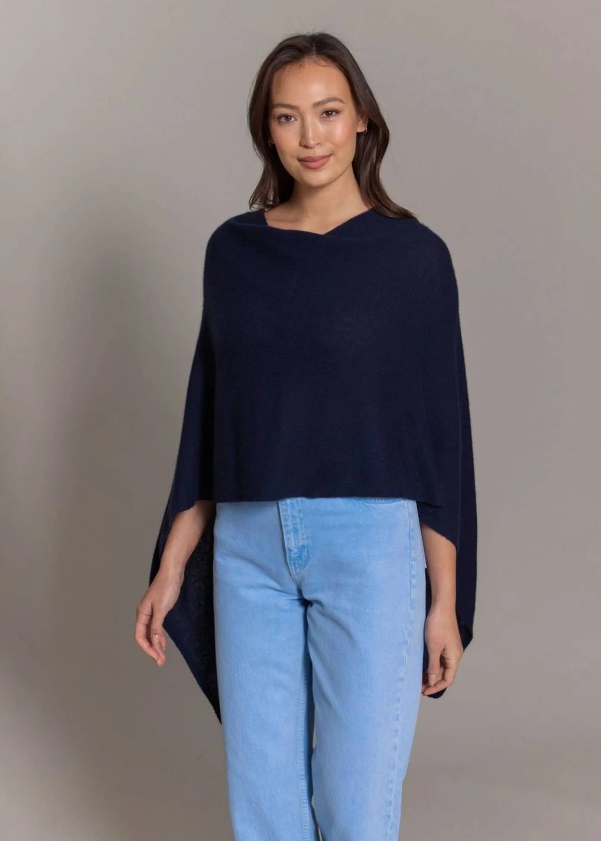 A woman modeling a navy blue knit poncho sweater paired with light blue jeans, standing against a neutral gray studio background.