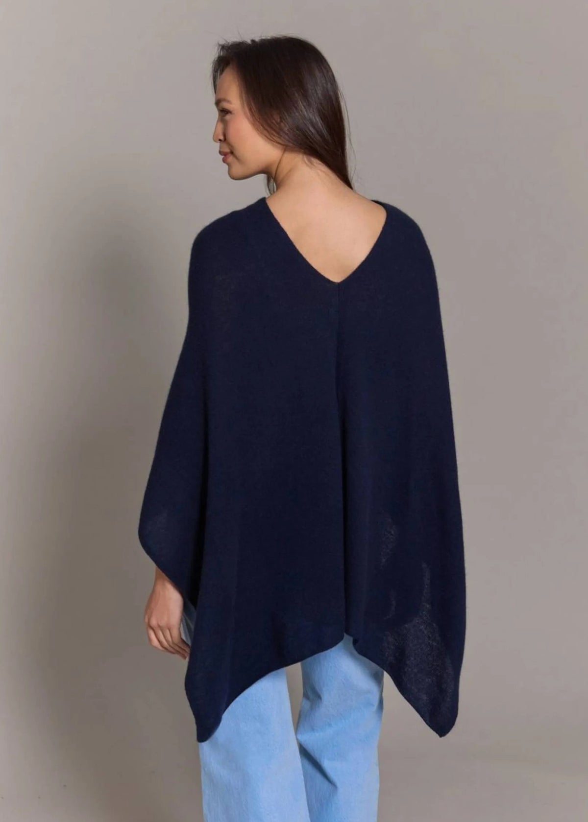 A woman shown from the back wearing a dark navy blue knit poncho-style top with a V-neck and light blue jeans, standing against a plain light gray studio background.