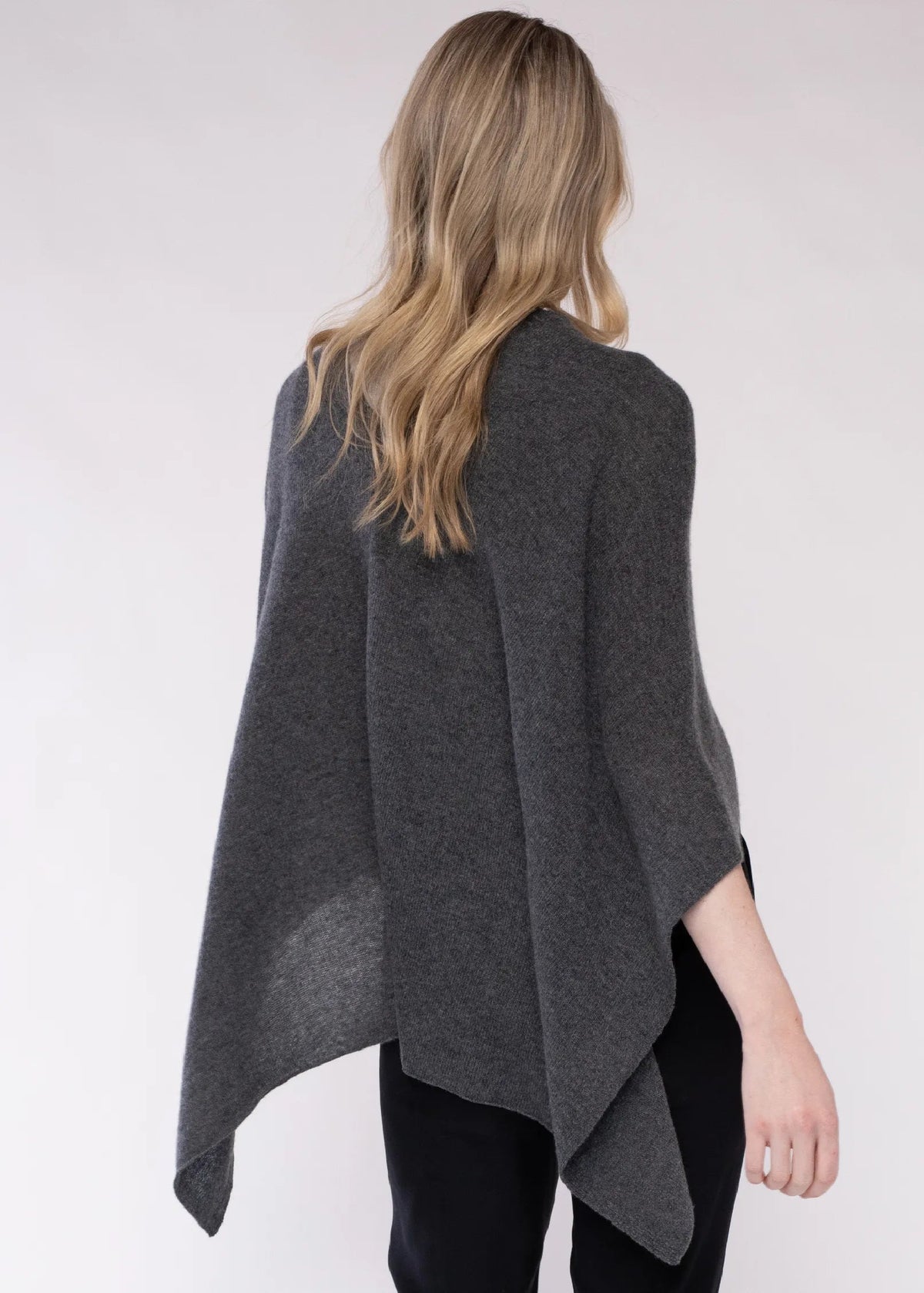 A woman viewed from behind wearing a dark gray open-front poncho or cape over black pants, against a plain light gray studio background.