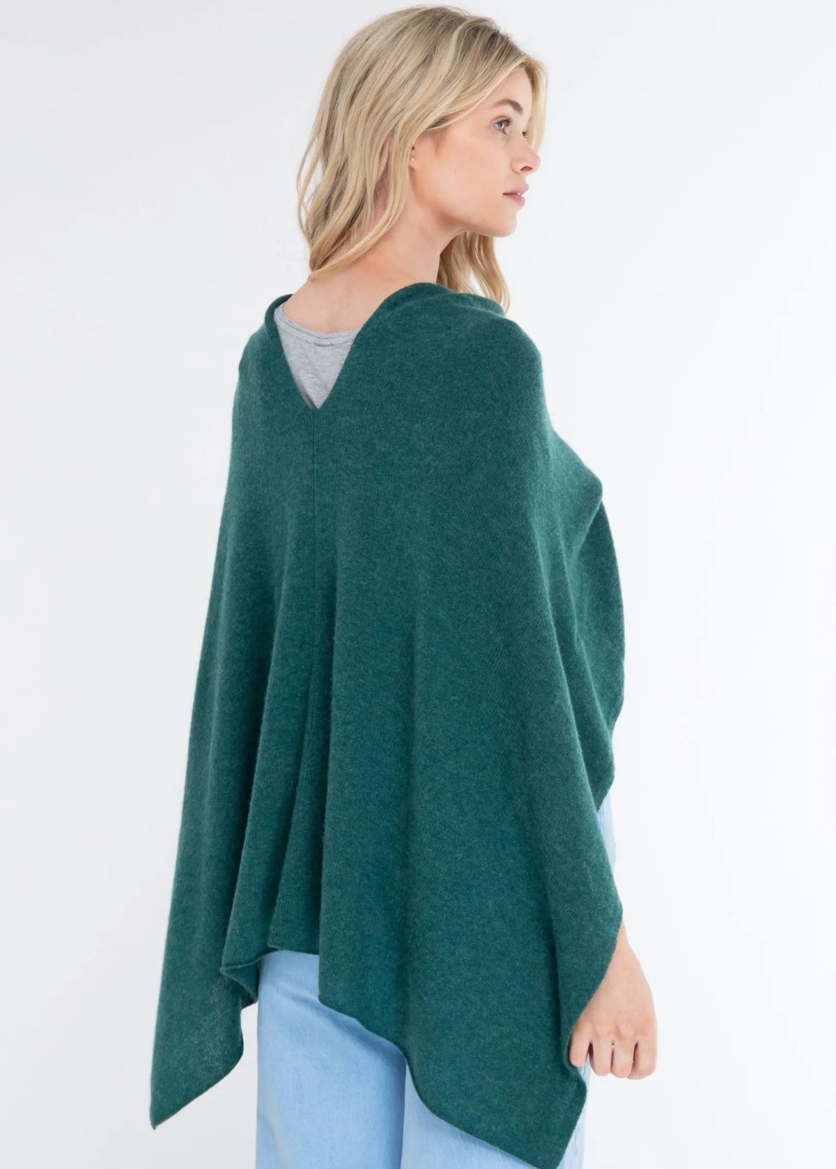 A woman wearing a dark green open-front poncho or cape over a gray top and light blue jeans, photographed against a plain white studio background.