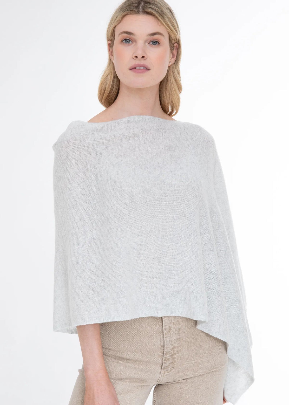 A woman modeling a light gray, semi-sheer knit poncho top with beige pants against a plain white studio background.