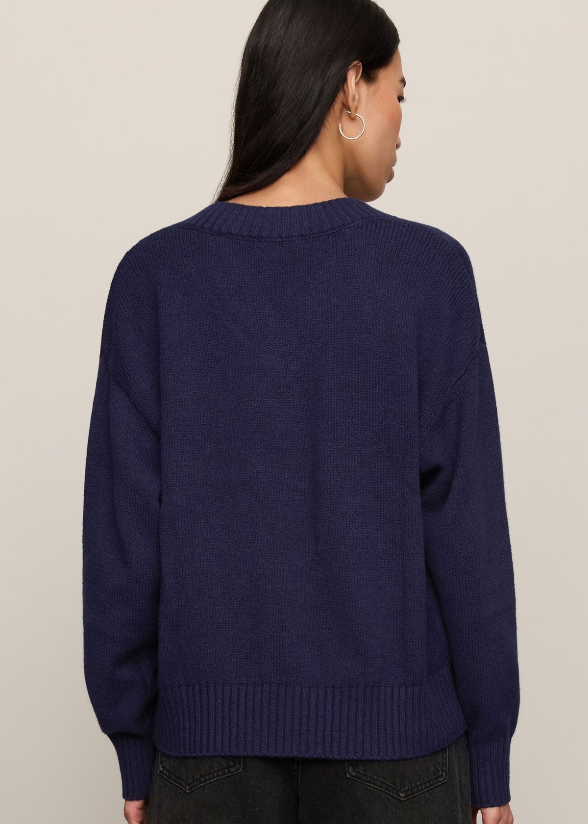 A woman shown from the back wearing a navy blue knit sweater and dark jeans against a plain light beige studio background.