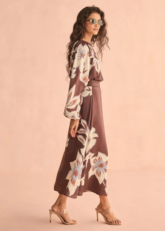 A woman modeling a brown maxi dress with a large cream, white, and light blue floral print, styled with clear aviator sunglasses and beige high-heeled sandals against a solid peach-pink studio background.