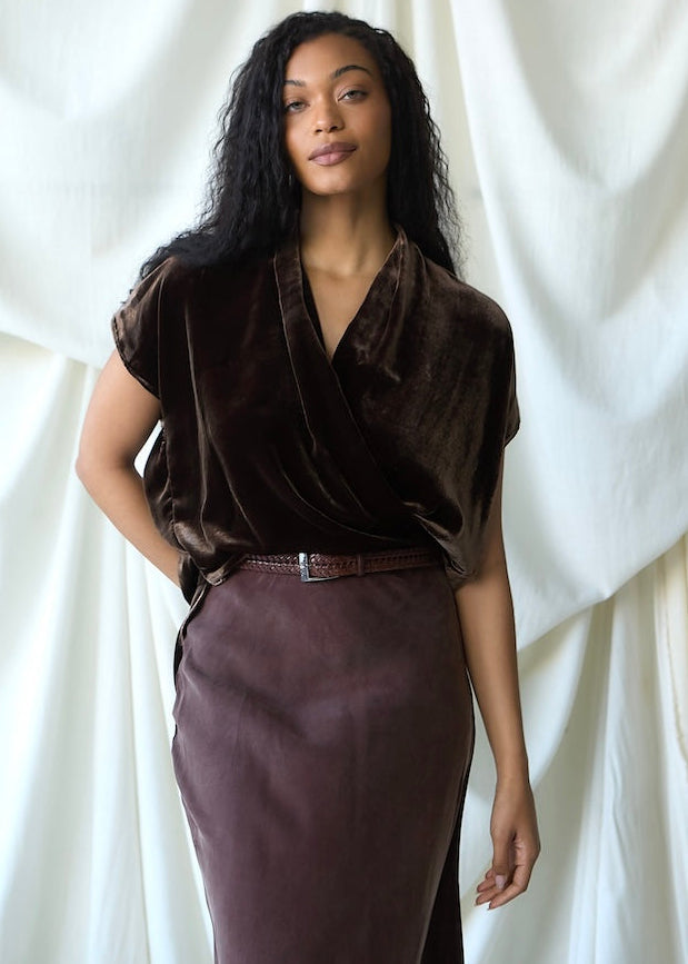 A woman modeling a dark brown velvet short-sleeve wrap top and a matching mauve-purple skirt, standing against a draped white fabric backdrop.