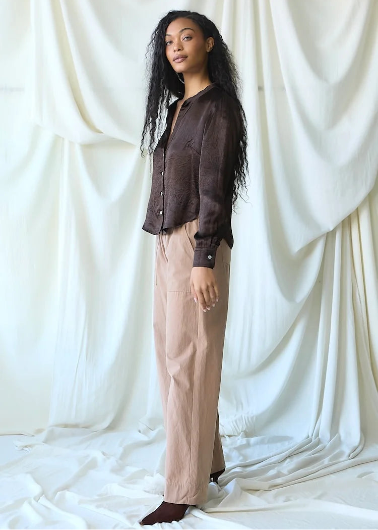 A woman stands in front of a draped white fabric backdrop, wearing a dark brown sheer button-up shirt and light beige wide-leg trousers.