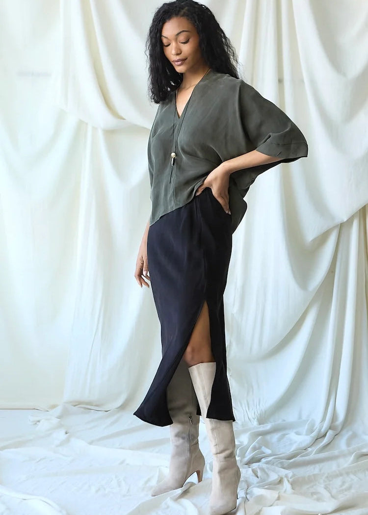 A woman models a two-piece outfit consisting of an olive green draped top and a black high-slit skirt, paired with off-white knee-high boots, set against a draped white fabric backdrop.