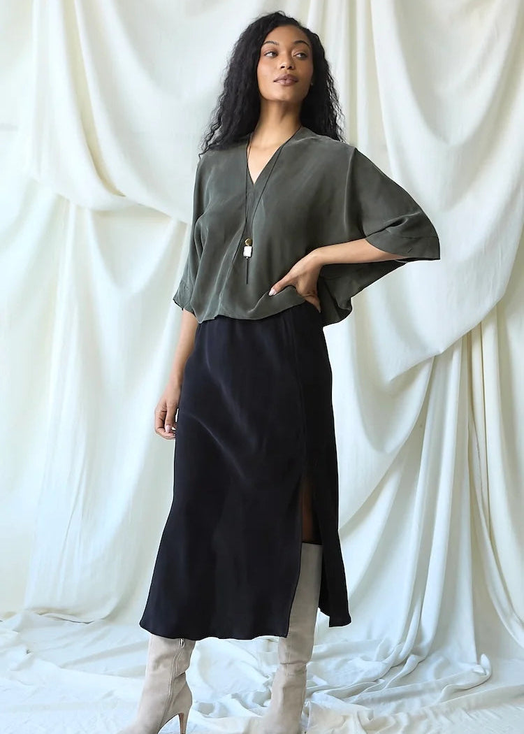 A woman models a dark olive green short-sleeve blouse with a V-neck and black midi skirt, paired with beige heeled boots, set against a draped white fabric backdrop.