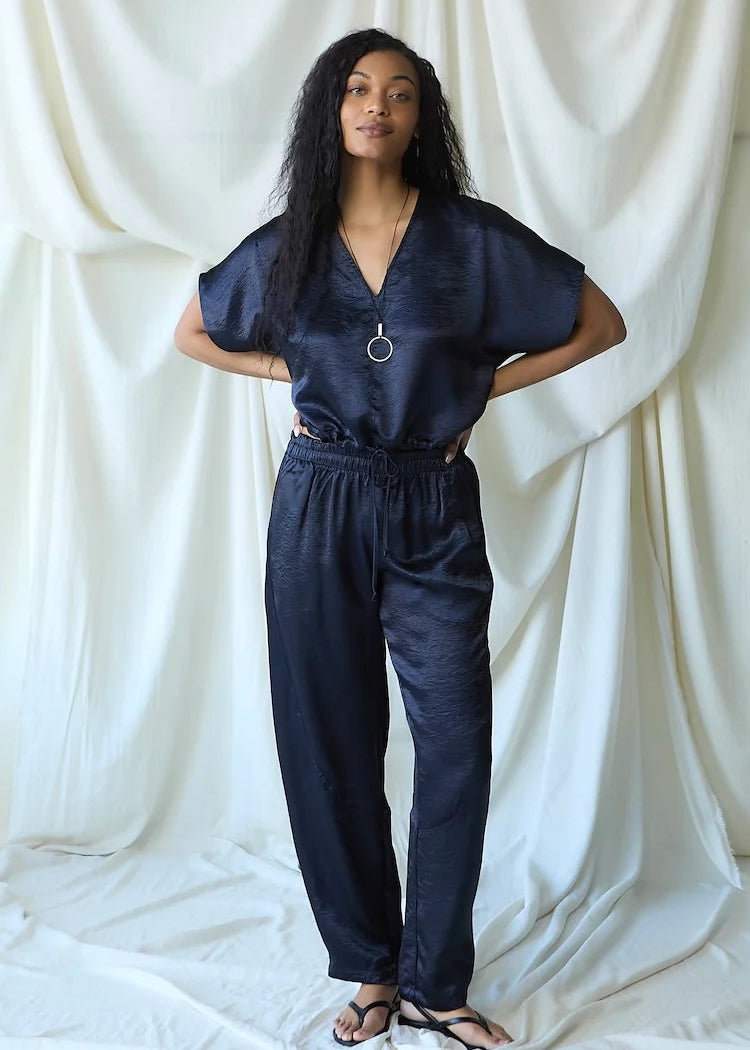 A woman modeling a dark navy blue two-piece set consisting of a short-sleeve V-neck top and matching drawstring pants, standing against a draped white fabric backdrop.