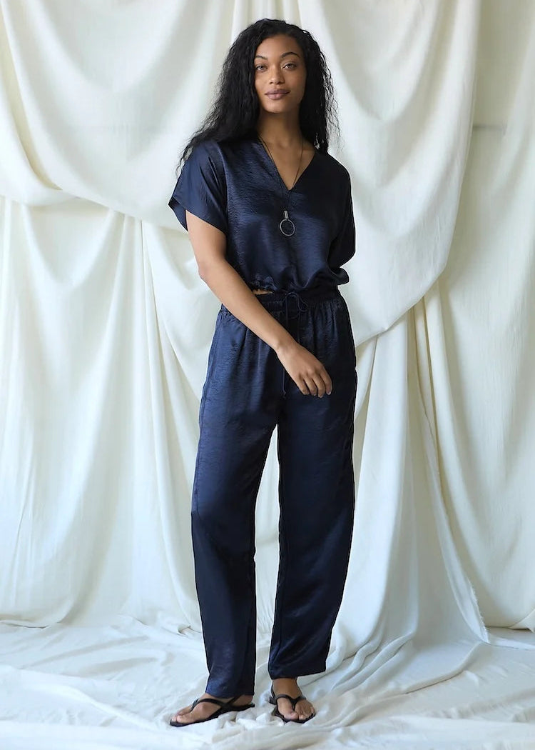 A woman modeling a dark navy blue satin jumpsuit with a V-neck, short sleeves, and a drawstring waist, standing against a draped white fabric backdrop.