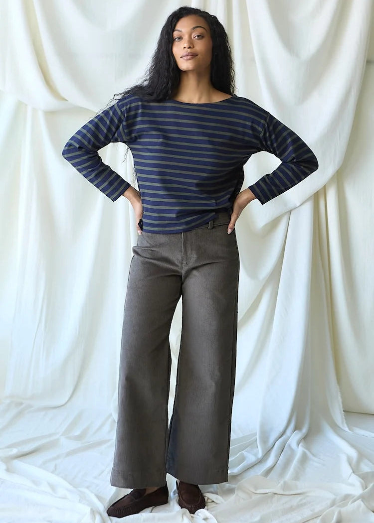 A woman stands with hands on hips wearing a navy blue and gray striped long-sleeve boatneck top with wide-leg gray pants, posed in front of a draped white fabric backdrop.