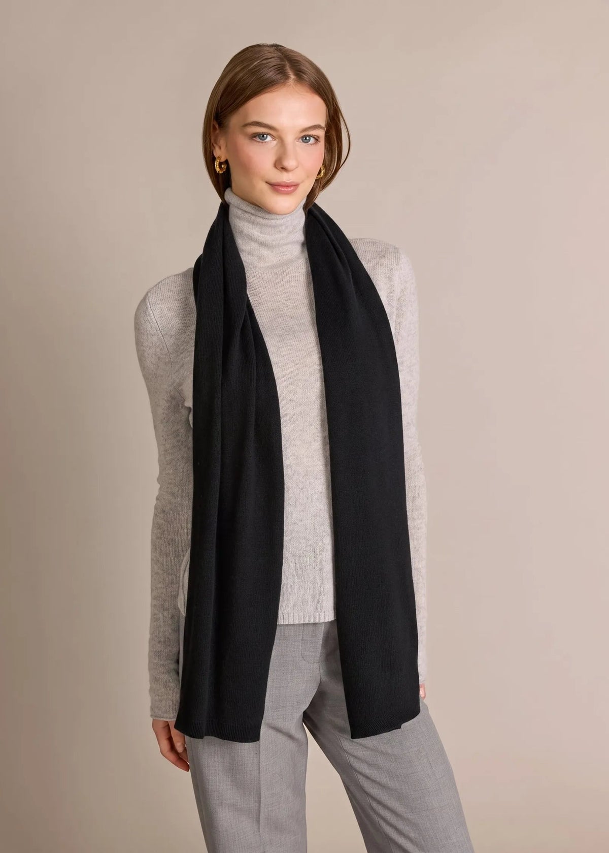 A woman modeling a long black scarf wrapped around her neck over a light gray turtleneck sweater and gray pants, set against a plain beige studio background.