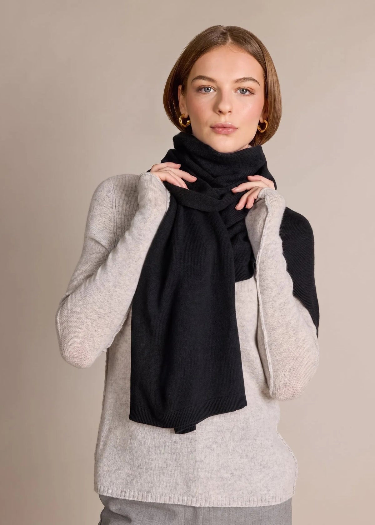 A woman modeling a black knit scarf wrapped around her neck over a light gray sweater, against a solid beige studio background.
