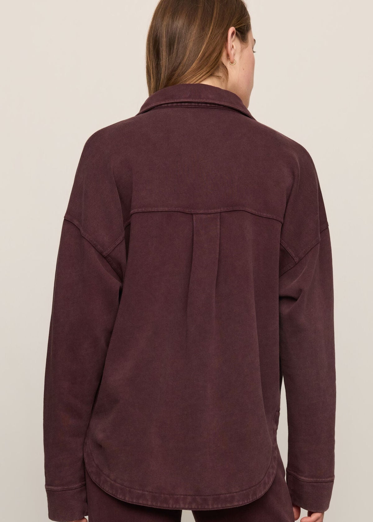 A woman viewed from the back wearing a dark plum button-up shirt and matching pants against a plain light beige studio background.