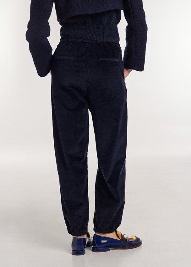 A person shown from the back wearing dark navy corduroy trousers, a matching dark knit top, and navy loafers with gold detailing, against a plain light gray studio background.