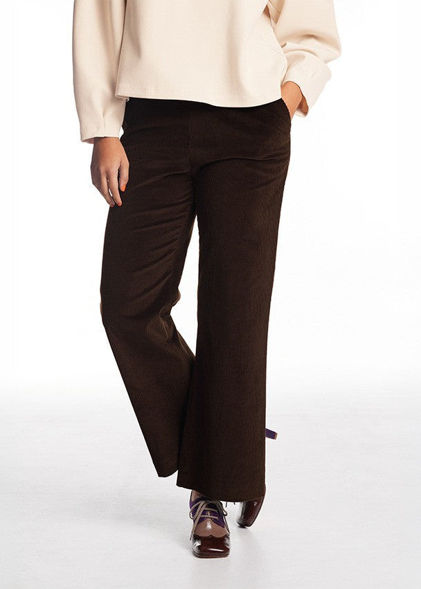 A model wearing dark brown wide-leg corduroy trousers, paired with a cream blouse and brown lace-up shoes, shown from the waist down against a white studio background.