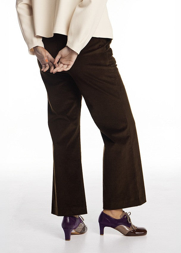 A person wearing dark brown wide-leg trousers, a cream-colored top, and purple high-heeled shoes, shown from the waist down against a white studio background.