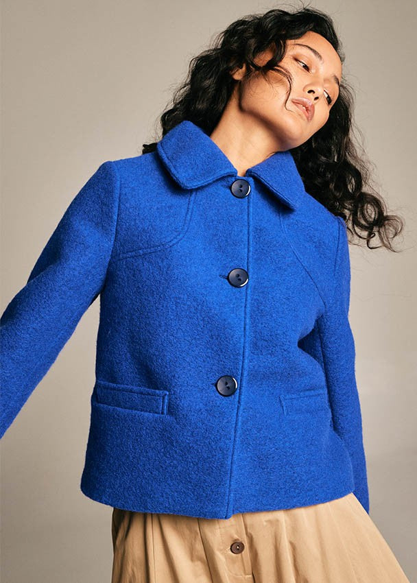A woman wearing a bright royal blue wool-blend jacket with a collar, three dark buttons, and front flap pockets, paired with a beige skirt, photographed against a plain warm gray studio background.