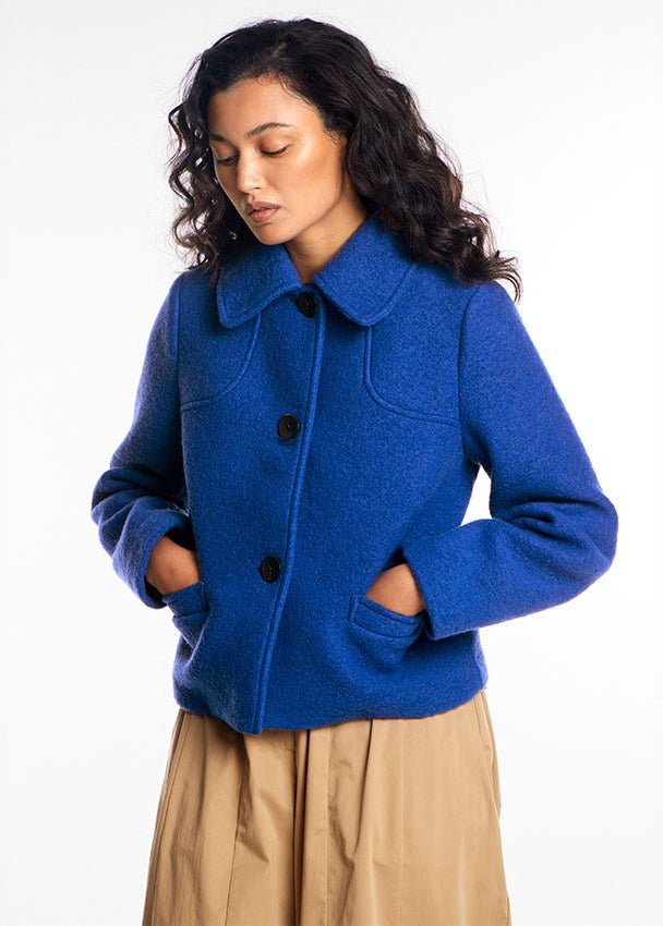 A woman wearing a bright royal blue wool-blend jacket with a wide collar, black buttons, and front patch pockets, paired with beige trousers, photographed against a clean white studio background.
