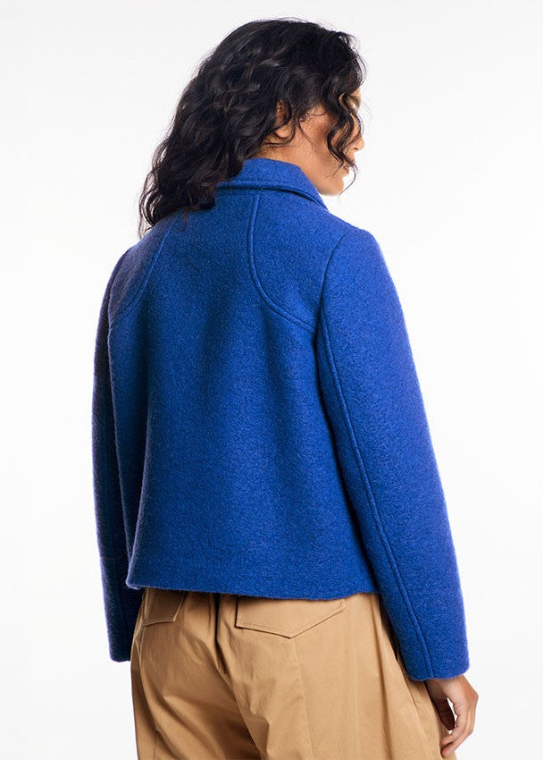 A woman shown from the back wearing a cropped, vibrant royal blue wool-blend jacket and tan trousers, photographed against a clean white studio background.