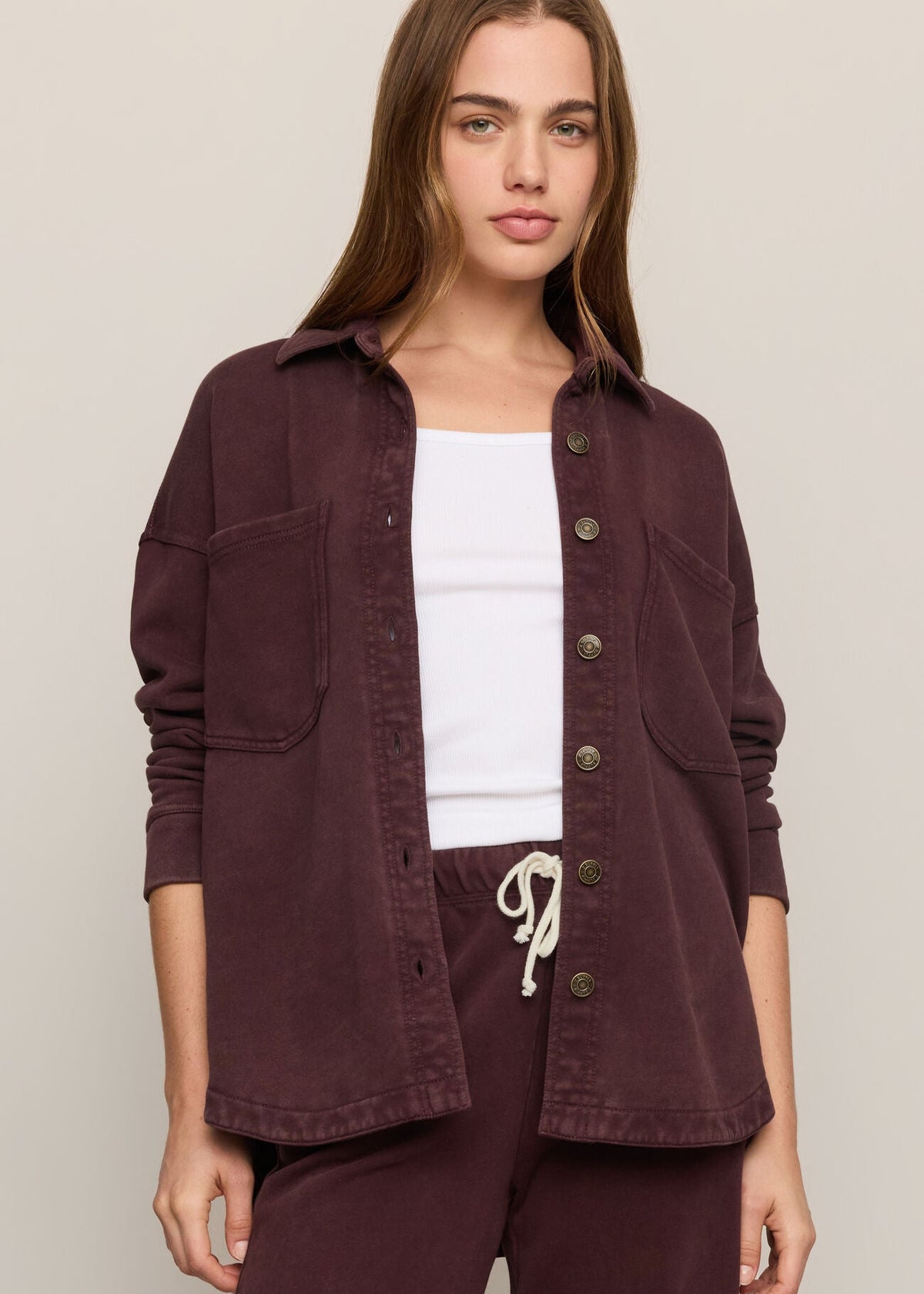 A woman modeling a dark plum button-up overshirt with front pockets, worn open over a white tank top and matching dark pants, against a plain beige studio background.