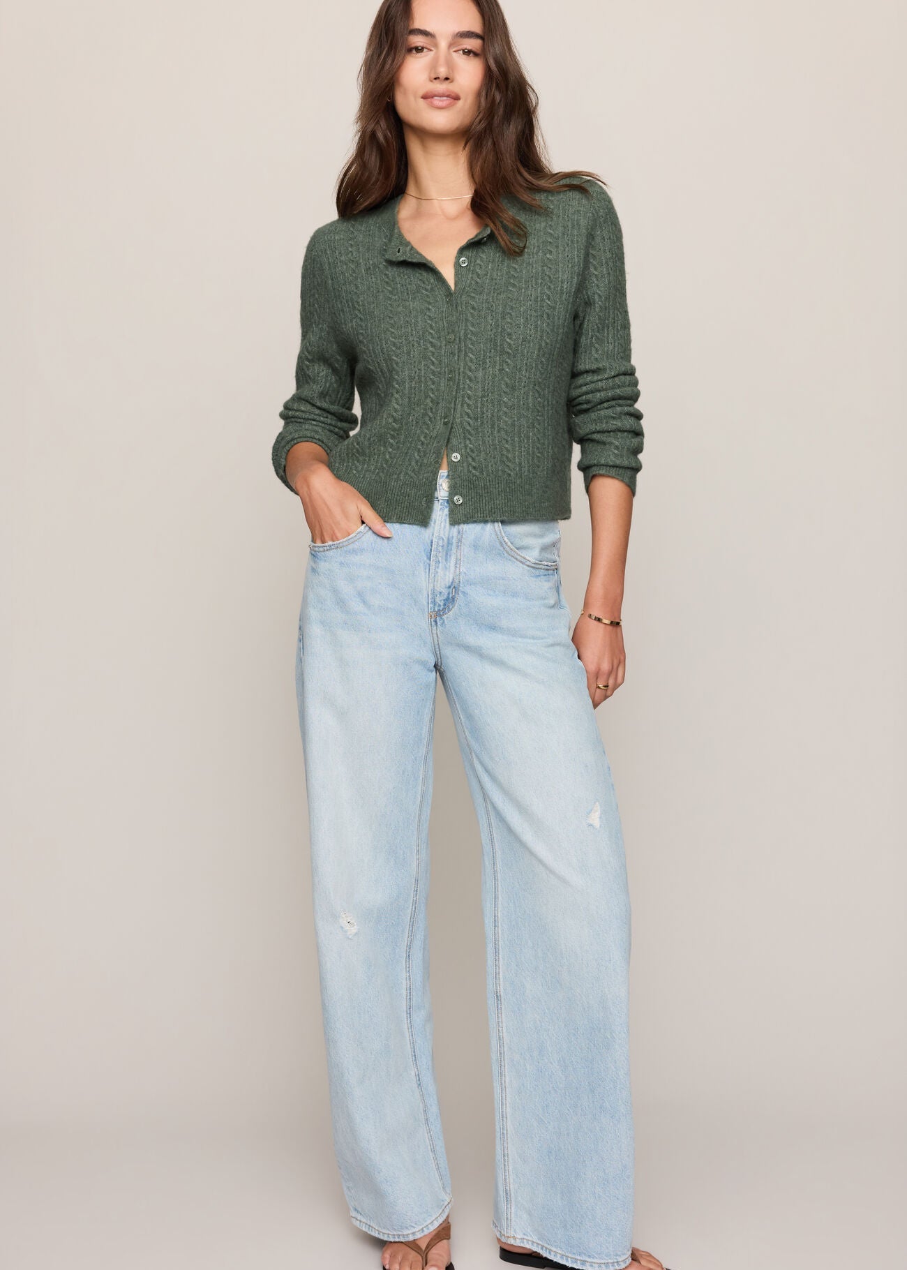 A woman modeling a dark green textured knit cardigan and light-wash wide-leg jeans against a solid beige studio background.