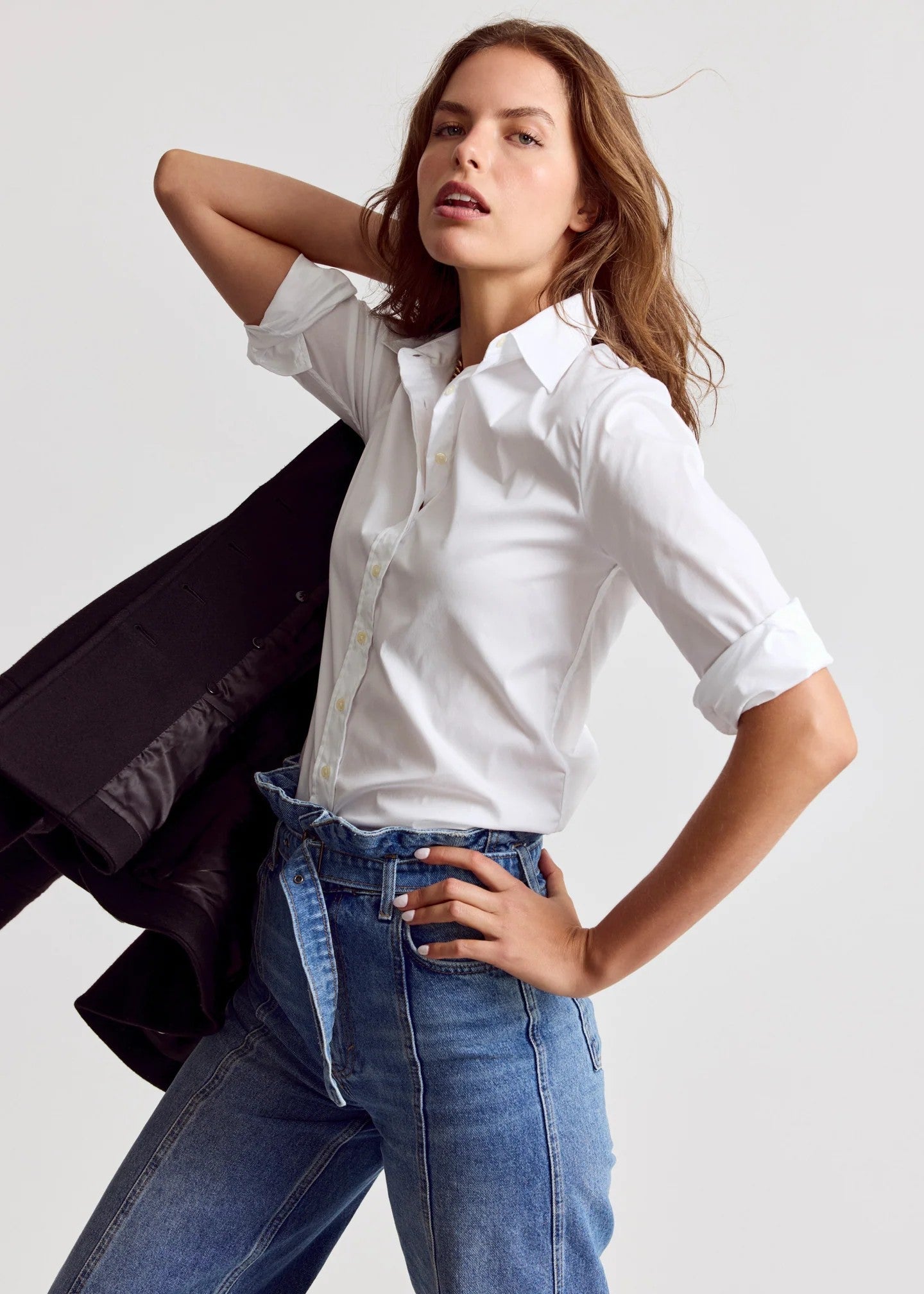 A woman posing in a white short-sleeve button-up shirt, blue jeans, and a black blazer against a light gray studio background.