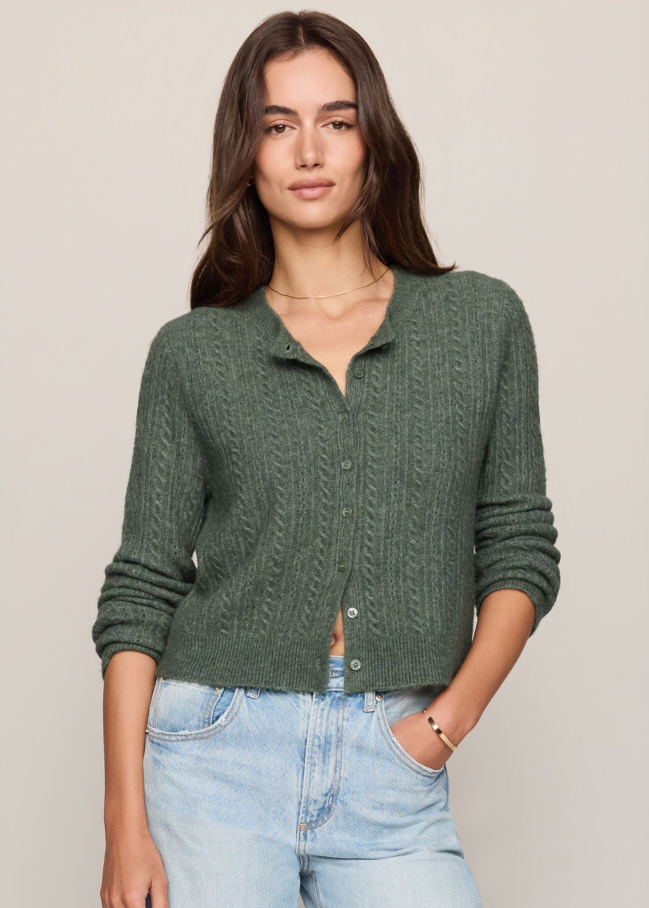 A woman modeling a dark green cable-knit button-up cardigan sweater paired with light-wash blue jeans, set against a plain beige studio background.
