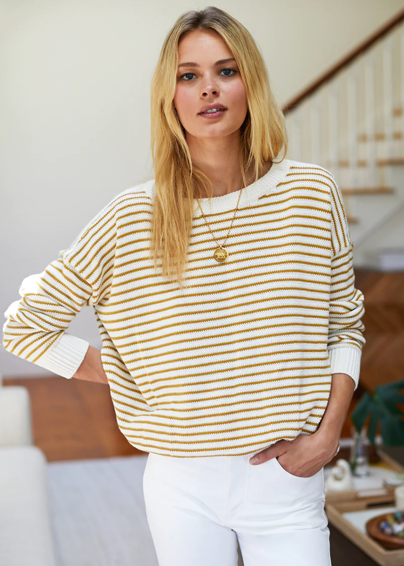 Woman wearing a yellow and white striped sweater and white pants indoors.