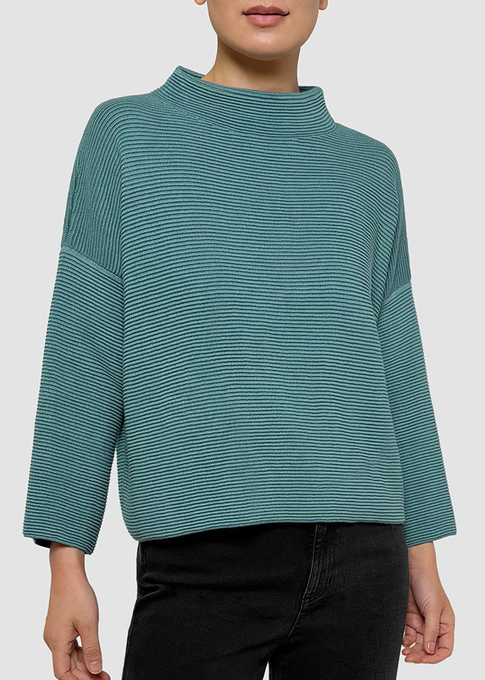 A woman wearing a teal-green long-sleeve mock neck knit top with thin horizontal stripes, paired with black pants, against a light gray studio background.