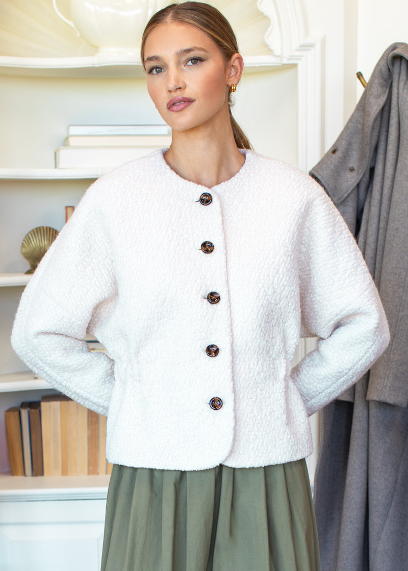 Woman wearing a white textured jacket with brown buttons in a room with shelves and a coat.