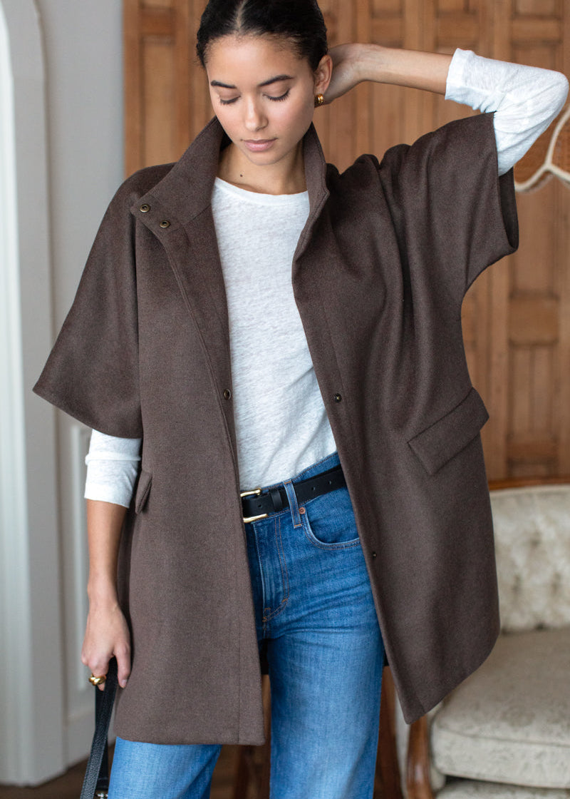 A woman wearing a brown open-front cape jacket over a white long-sleeve top and blue jeans, standing indoors in a room with wood paneling and a tufted chair.